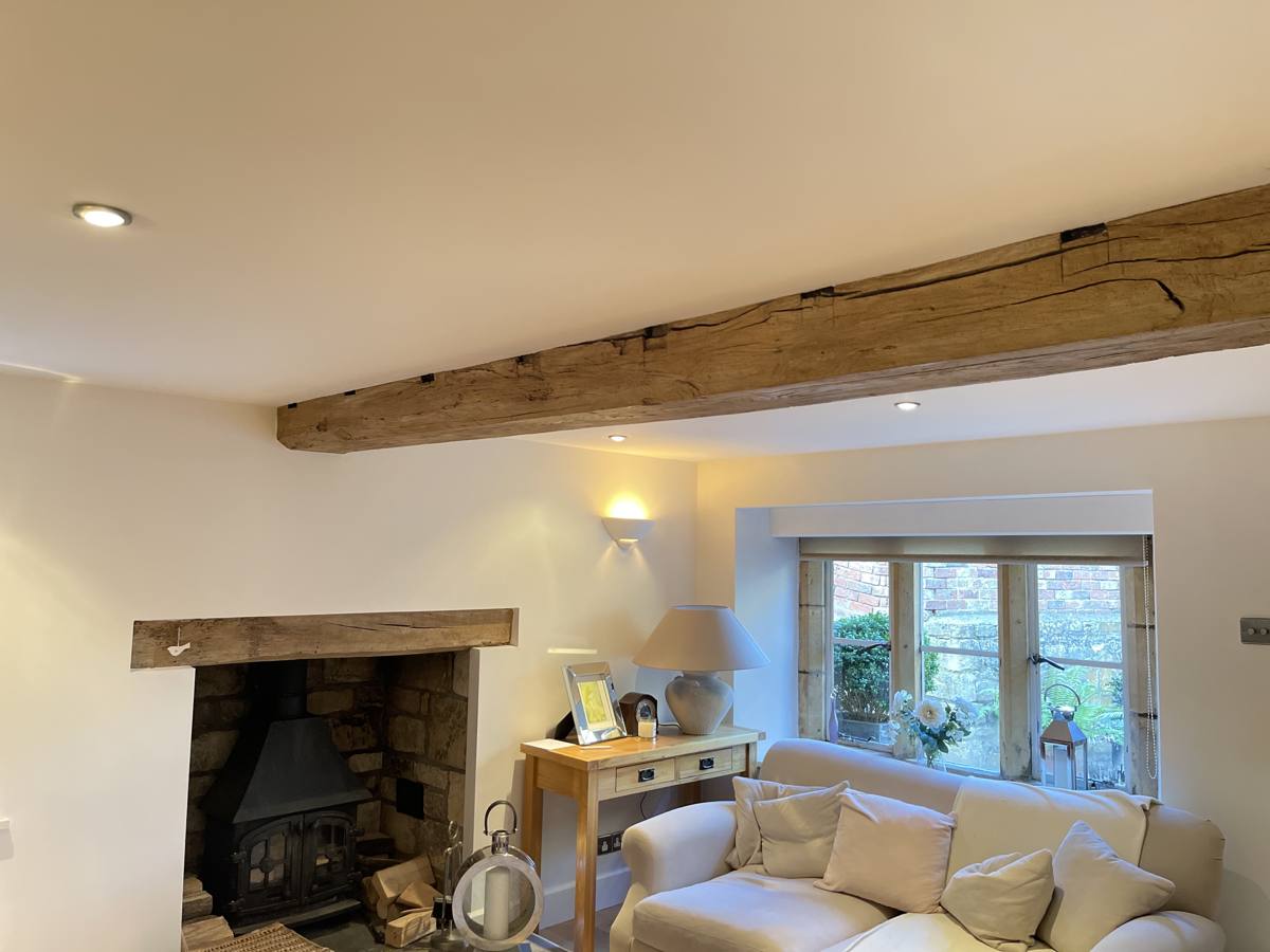 Restored oak ceiling beams in a Cotswolds cottage sitting room with a stone inglenook and wood-burning stove